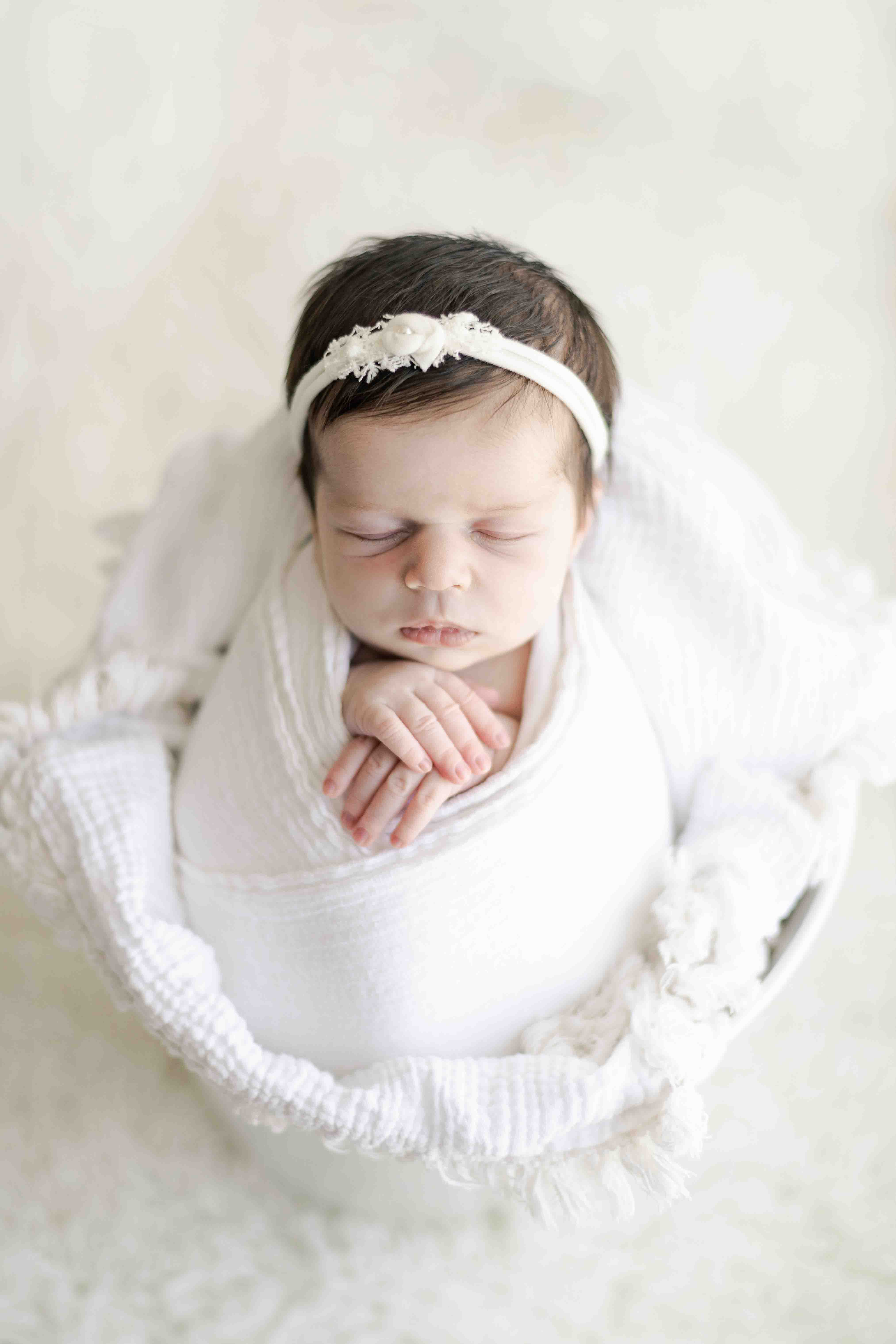 Newborn baby girl photography – peacefully posed in a white bucket, hands gently folded, wearing a delicate headband with a tiny bow. A timeless and pure newborn portrait.