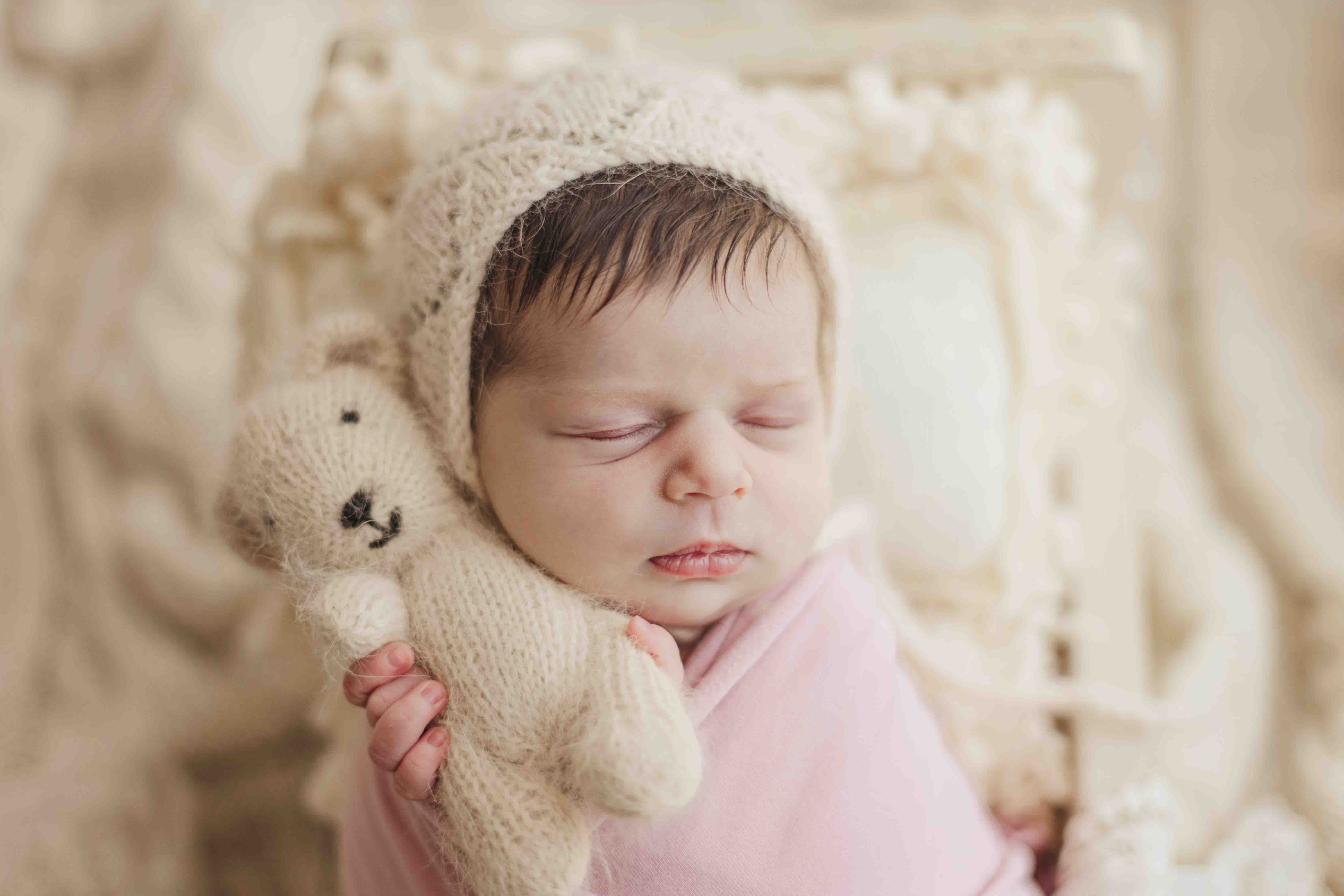 Sandra Hext Photography – Newborn baby girl photography – snuggled in soft neutral beige tones, holding a tiny bear, wrapped in a delicate mauve wrap. A warm and timeless portrait capturing pure sweetness.